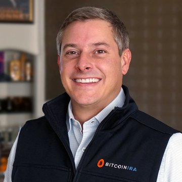 A professional headshot of a smiling middle-aged man wearing a dark vest over a light blue shirt, featuring an orange "BITCOINIRA" logo on his chest.
