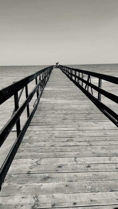 A black and white photograph of a long wooden pier extending out into calm ocean waters toward the horizon under a hazy sky.