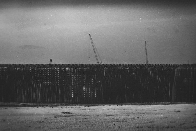 A moody black-and-white photograph of a construction site featuring a long barrier or wall of vertical panels with two tall cranes silhouetted against a hazy sky.