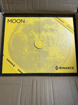 A Binance-branded 'MOON' yellow coin or medal inside a black case, featuring a moon surface design and the Binance logo in the bottom right corner.