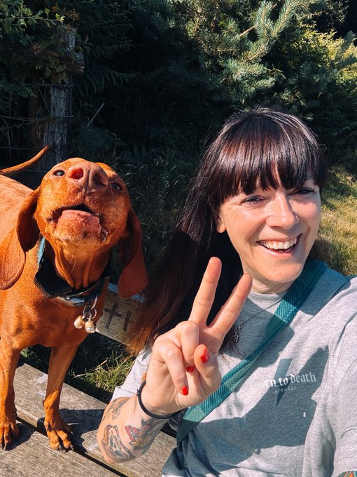 A smiling woman with dark hair makes a peace sign next to a reddish-brown dog (Vizsla) with its mouth open, in an outdoor setting with trees.