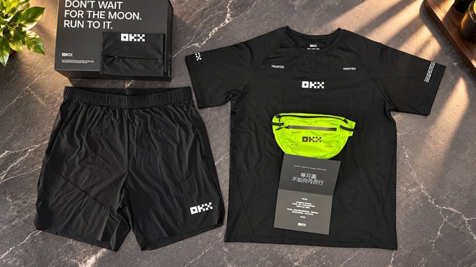 An OKX branded fitness kit laid out on a dark marble surface, containing black shorts, a black t-shirt, a yellow waist bag, and a box with the slogan 'DON'T WAIT FOR THE MOON. RUN TO IT.'