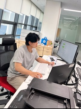 A young man sits at a desk in a bright office working on two monitors and a laptop, with a keyboard and printer visible on the workspace.