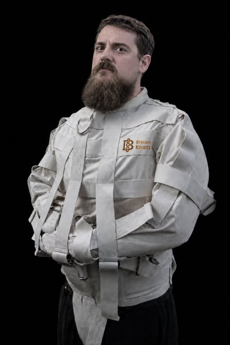 A promotional photo of a bearded man wearing a straitjacket with a 'Bitcoin Knots' logo embroidered on it.