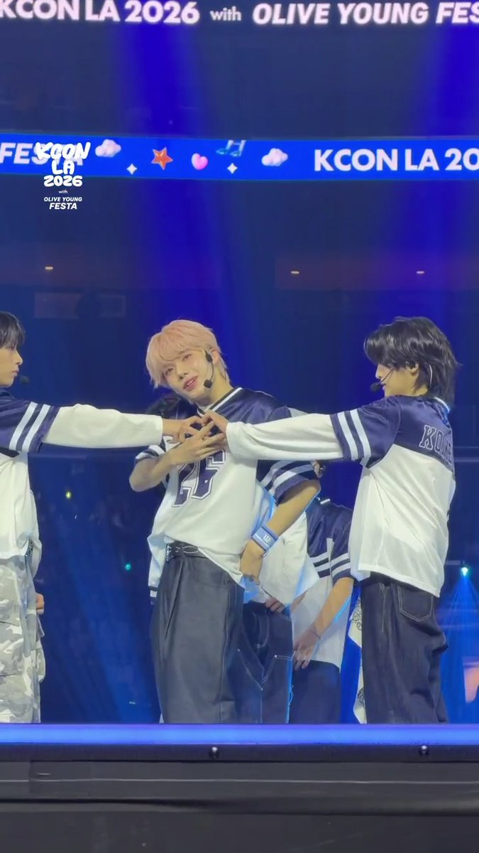 Performers in matching white and navy baseball-style jerseys form a heart shape with their hands on stage at KCON LA 2026 with OLIVE YOUNG FESTA, as shown by the banners and stage branding visible above them.