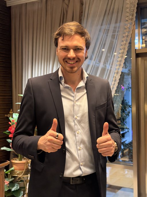 A smiling man in a dark blazer and white dress shirt gives two thumbs up, appearing dapper and professional in his new "suitcoin" attire, fitting the caption about his transition into a suit-wearing executive role at Big Bitcoin.