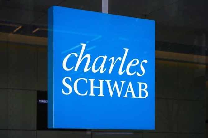 An illuminated blue square sign displaying the 'charles SCHWAB' logo in white text, mounted on the exterior of a glass building.