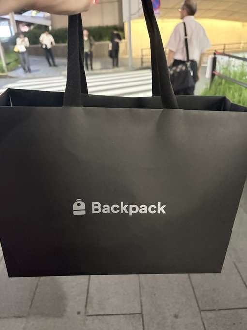 A hand holding a black tote bag with the 'Backpack' exchange logo, taken outside on a street with pedestrians in the background.