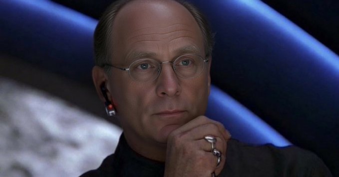 The image shows a bald man with glasses, wearing a black shirt and ring on his finger, sitting or standing by himself against a blue background that has text. The caption at the bottom of the image talks about a new cryptocurrency called XLM outperforming XRP in terms of market value.