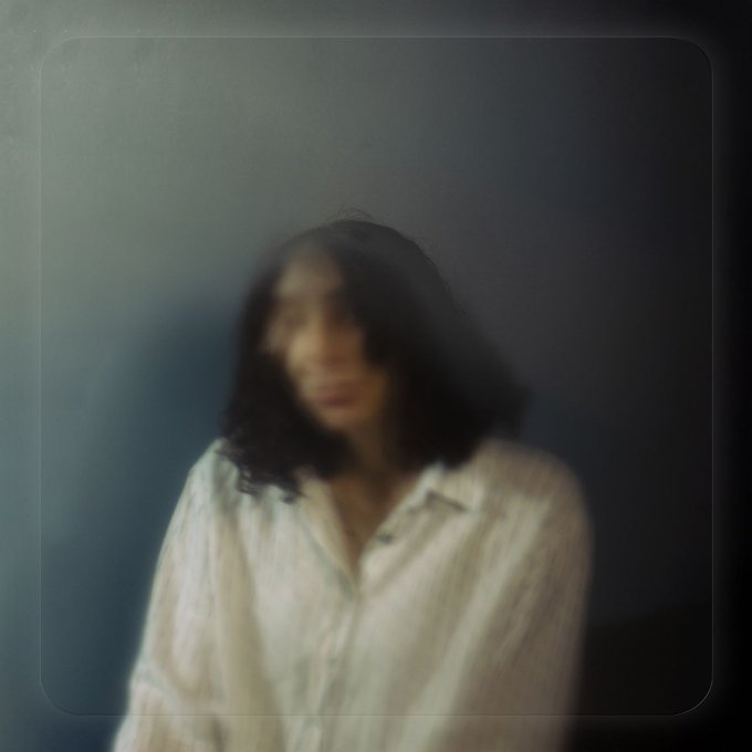 A heavily blurred, moody portrait of a woman with dark hair wearing a white striped button-up shirt, set against a dim, dark background with a vintage border effect.