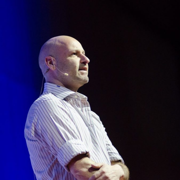 A bald man wearing a striped button-down shirt and a headset microphone speaks on stage, illuminated by blue lighting, appearing to deliver a talk on cryptocurrency and blockchain technology. The image likely depicts Joseph Lubin, the Ethereum co-founder mentioned in the post, as he discusses Ethereum's low Layer 1 fees and the future of onchain companies.