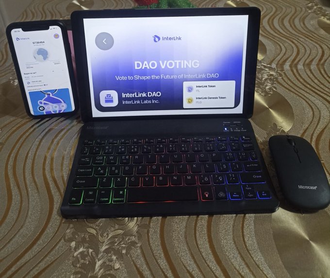 A photograph of a tablet displaying 'InterLink DAO VOTING' for 'InterLink DAO' with 'InterLink Labs Inc.', placed next to a smartphone showing a digital wallet/portfolio and a black wireless mouse, all resting on a golden patterned tablecloth.