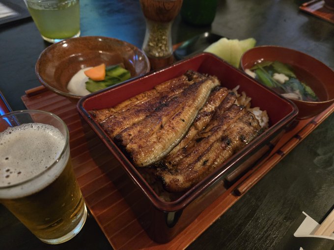 A Japanese meal featuring a grilled eel (unagi) rice bowl in a red lacquer box, accompanied by side dishes and a glass of beer on a wooden tray.