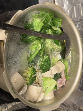 A metal pot containing a soup with noodles, tofu, broccoli, bok choy, and meat slices, with a ladle inside.