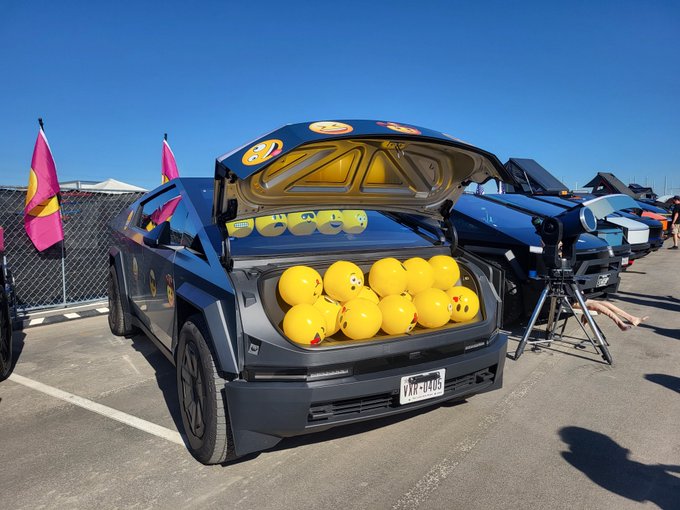 A black Tesla Cybertruck with its rear hatch open, revealing a trunk filled with yellow smiley-face balloons, parked near a chain-link fence with pink flags.
