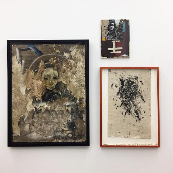 Three framed artworks hang on a white gallery wall, featuring two weathered, distressed portrait pieces with ghostly figures and a small mixed-media collage with a cross-like shape.