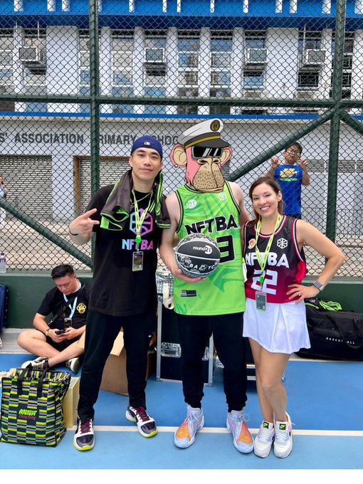 Two people pose on a basketball court flanking a mascot character wearing a green "NFTBA" jersey and sailor hat, with the mascot holding a basketball.