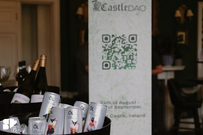A bottle of wine and several cans of 'ÖL' cider sit in a bucket in front of a standee for 'CastleDAO' featuring a QR code and event dates in Castle, Ireland.