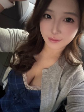 A selfie of a young woman with long wavy hair, wearing a denim dress and gray cardigan, smiling softly at the camera.