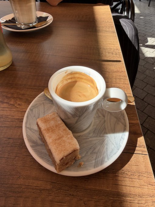 A ceramic cup of espresso with rich, golden crema sits on a patterned saucer alongside a small piece of cake, placed on a sunlit wooden café table. In the background, another coffee drink is partially visible, suggesting a relaxed café moment centered around the bold, concentrated shot of espresso.