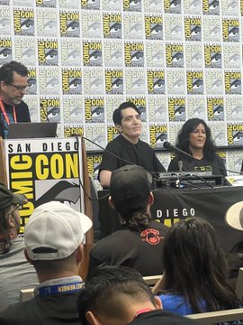A panel at San Diego Comic-Con with attendees seated at a table in front of a Comic-Con backdrop, with audience members visible in the foreground.
