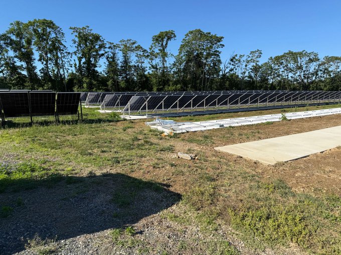 The image shows an empty field with several rows of solar panels installed across its surface, indicating that this area is part of a solar farm. The photo also features a small patch of grass near the edge of the field, adding some natural elements to the scene. The caption provided in the image seems unrelated and does not provide any context or information about the solar farm itself.