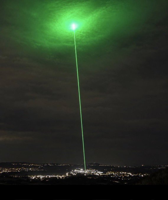 A nighttime photograph of a brilliant green laser beam shooting vertically into the sky from an illuminated city below, creating a glowing halo in the clouds above.