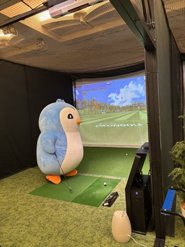 A large blue penguin mascot stands on a green mat inside an indoor golf simulator, facing a screen displaying a golf course with the 'OKONGOLF' logo.
