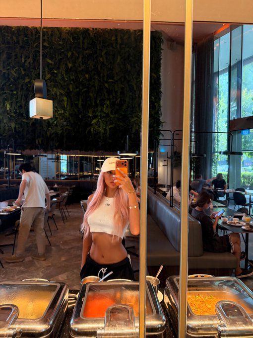 A young woman with pink hair takes a mirror selfie in a restaurant with a green plant wall, showing food serving trays in the foreground.
