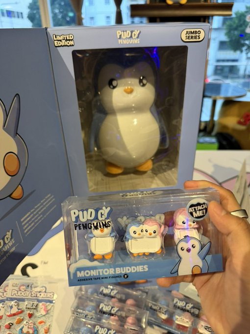 A hand holding a packaged set of Pudgy Penguins "Monitor Buddies" adhesive tape mini figures, with a larger Pudgy Penguins Jumbo Series box displayed in the background.