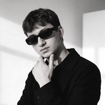 A black and white portrait of a young man wearing dark sunglasses and a black shirt, posing with his hand near his chin.