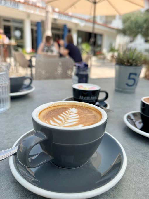 A close-up photograph of a cappuccino with latte art in a blue cup and saucer on an outdoor cafe table, with people sitting in the blurred background.