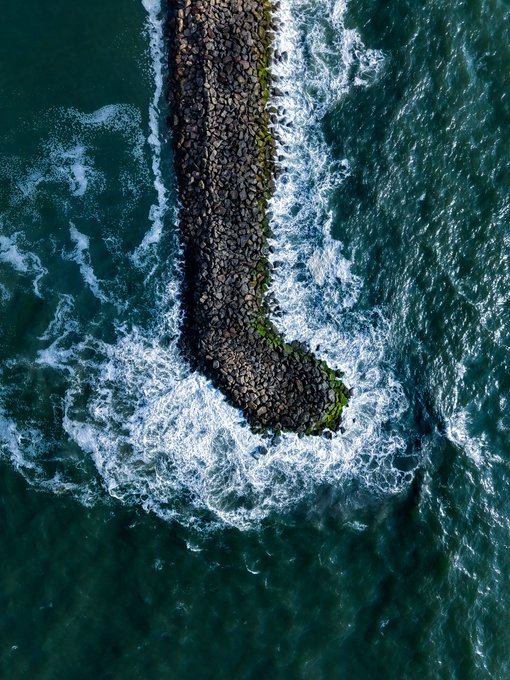 An aerial view captures a stone breakwater extending into the ocean, where white-capped waves crash against its rocky surface, creating a dynamic contrast between the deep teal water and the foaming surf. The image evokes the rhythmic, powerful motion of the sea—fitting imagery for a "Wave symphony" NFT mint being kicked off on TransientLabs.