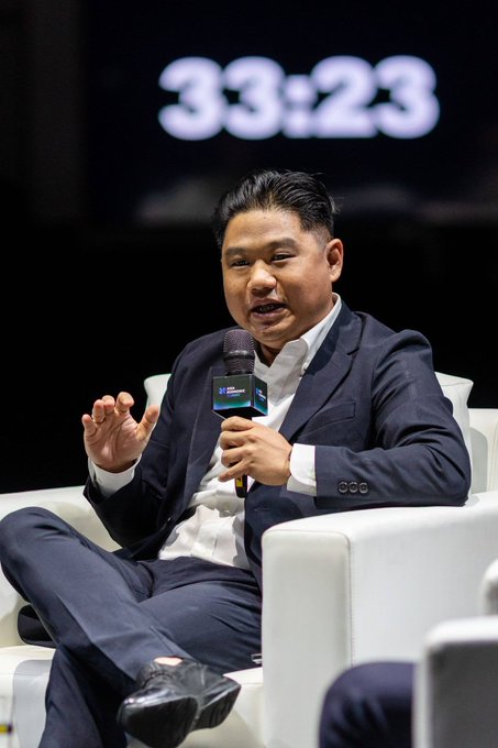 A man in a dark suit sits in a white chair on stage, speaking into a microphone while gesturing with his other hand, with a large timer reading "33:23" displayed behind him. The setting appears to be a panel discussion or interview, aligning with the post's introduction of Nael, CEO of IDRX, as a featured builder at an APAC-focused event.