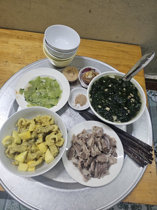 A top-down view of a home-cooked Chinese meal on a round tray with stacked bowls, featuring a chicken soup with greens, stir-fried chicken with potatoes, sliced boiled meat, sautéed cabbage, and a small dish of seasoning.