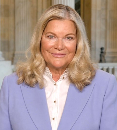 A blonde woman in a lavender blazer and white blouse smiles at the camera in what appears to be a formal government building setting with classical columns. The image accompanies a post about U.S. Senator Lummis proposing a plan to sell U.S. gold reserves to purchase 5% of all Bitcoin to address the $40 trillion national debt.