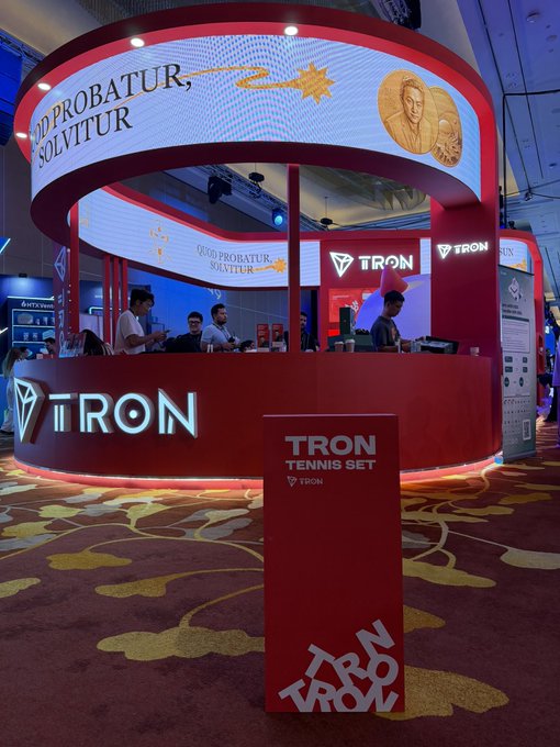 A circular TRON booth at an event features Latin text 'QUO PROBATUR, SOLVITUR', coin graphics with a face, and a red 'TRON TENNIS SET' stand labeled 'TURN UP 2024'.