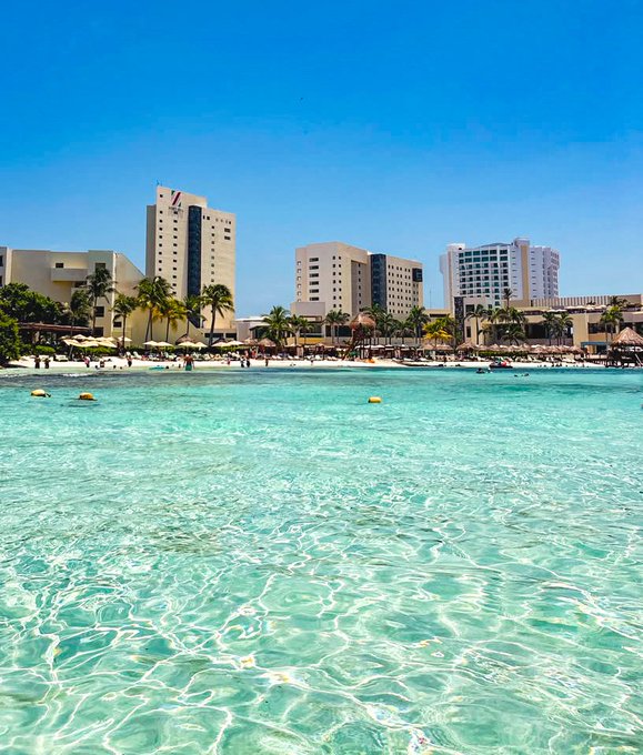 A bright tropical beach scene featuring crystal-clear turquoise water in the foreground, with a sandy beach lined with palm trees, lounge chairs, and tall white resort hotels in the background under a clear blue sky.