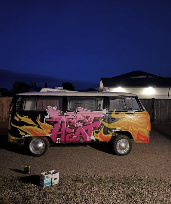A vintage Volkswagen van features a vibrant graffiti mural reading "BEAT THE HEAT" in bold pink lettering surrounded by yellow and orange flames, photographed at dusk. The post explains the artwork was painted for the Beat the Heat skate competition, with ramps built on top of the van for the event.
