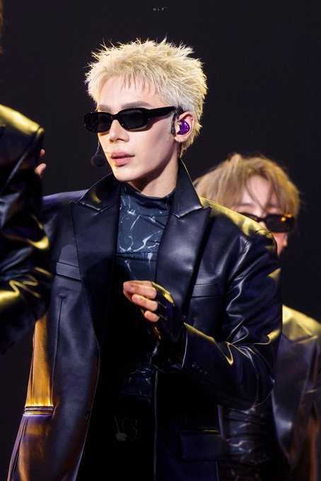 A performer with spiky blonde hair, black sunglasses, and a black leather blazer holds a microphone on stage, with another performer partially visible behind him.