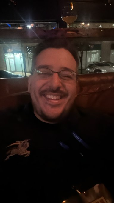 A selfie of a smiling man with glasses and a mustache wearing a black hoodie, taken at night outside with a car and streetlights in the background.