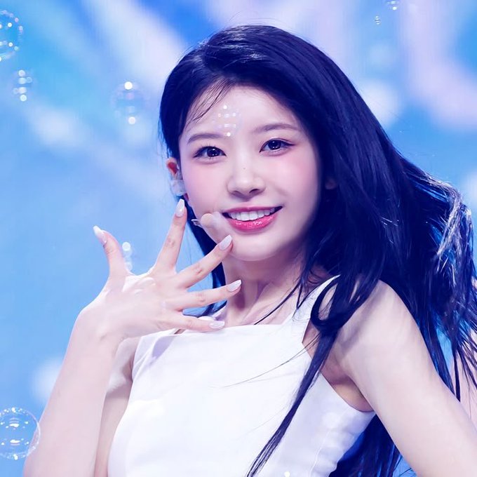 A young woman with long dark hair and decorative pearl-like facial gems smiles brightly while making a peace sign on a blue-lit stage, wearing a white sleeveless top and headset microphone—appearing to be a K-pop idol performing. Her cheerful, playful pose complements the post's lighthearted tone about missing out on an "Aurus VIP" upgrade, as she gestures toward the camera as if to reassure fans that a lottery or app upgrade will indeed be available.