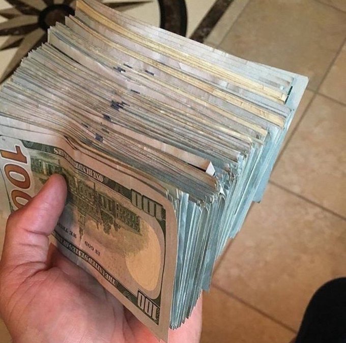 A hand grips a thick stack of $100 U.S. bills, fanned out to show the "100" denominations on the edges of the notes. The image visually represents the hustle and financial grind referenced in the caption.