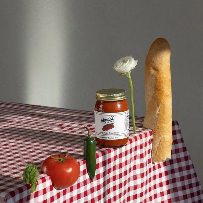 A still life on a red-and-white checkered tablecloth featuring a tomato, jalapeño, basil, a jar of Monti's Fireside sauce, a white flower, and a baguette bathed in dramatic side lighting.