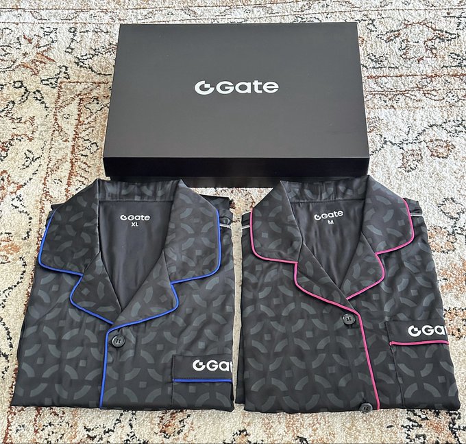 Two folded Gate.io branded pajamas (size XL with blue trim and size M with pink trim) displayed in front of a black Gate.io gift box on a patterned rug.