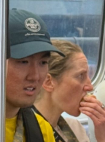 Two people sit on public transit, including an East Asian man in a dark green cap and a woman eating something in the background.