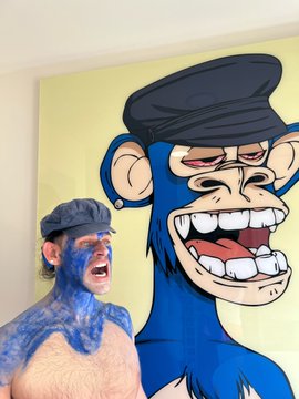 A shirtless man with blue paint on his face and wearing a blue headband stands next to a large painting of a blue cartoon monkey.