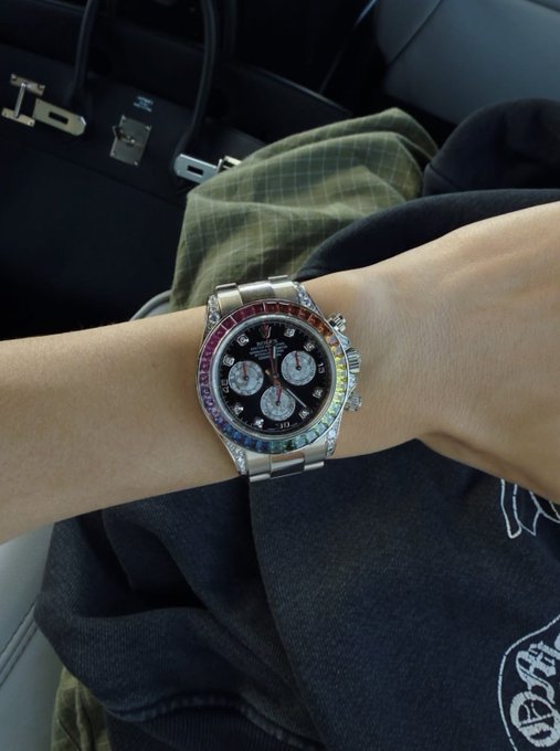 The image shows a wrist wearing a Rolex Daytona watch with a distinctive rainbow/sapphire bezel and black dial, set against what appears to be a car interior with a luxury bag visible in the background, reflecting a flex of wealth that complements the "$SOL" crypto and "drop your wallets" call to action.