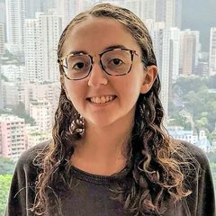 A selfie of a smiling young woman with curly brown hair and glasses, with a cityscape of high-rise buildings visible in the background.