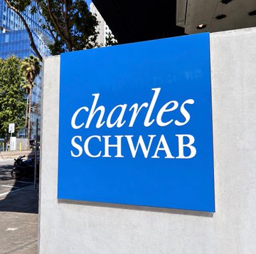 A blue "charles SCHWAB" sign is displayed prominently outside a building, with trees and a city skyline visible in the background, visually representing the financial giant mentioned in the announcement about their new Bitcoin and crypto trading services for eligible clients.